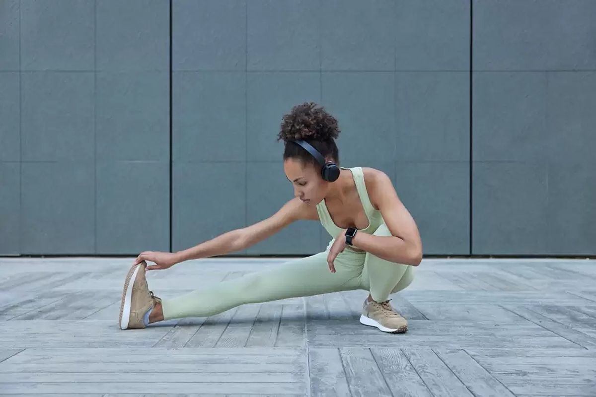 Horizontal shot of active slim woman stretches legs demonstrates her flexibility listens audio track in wireless headphones wears tracksuit.